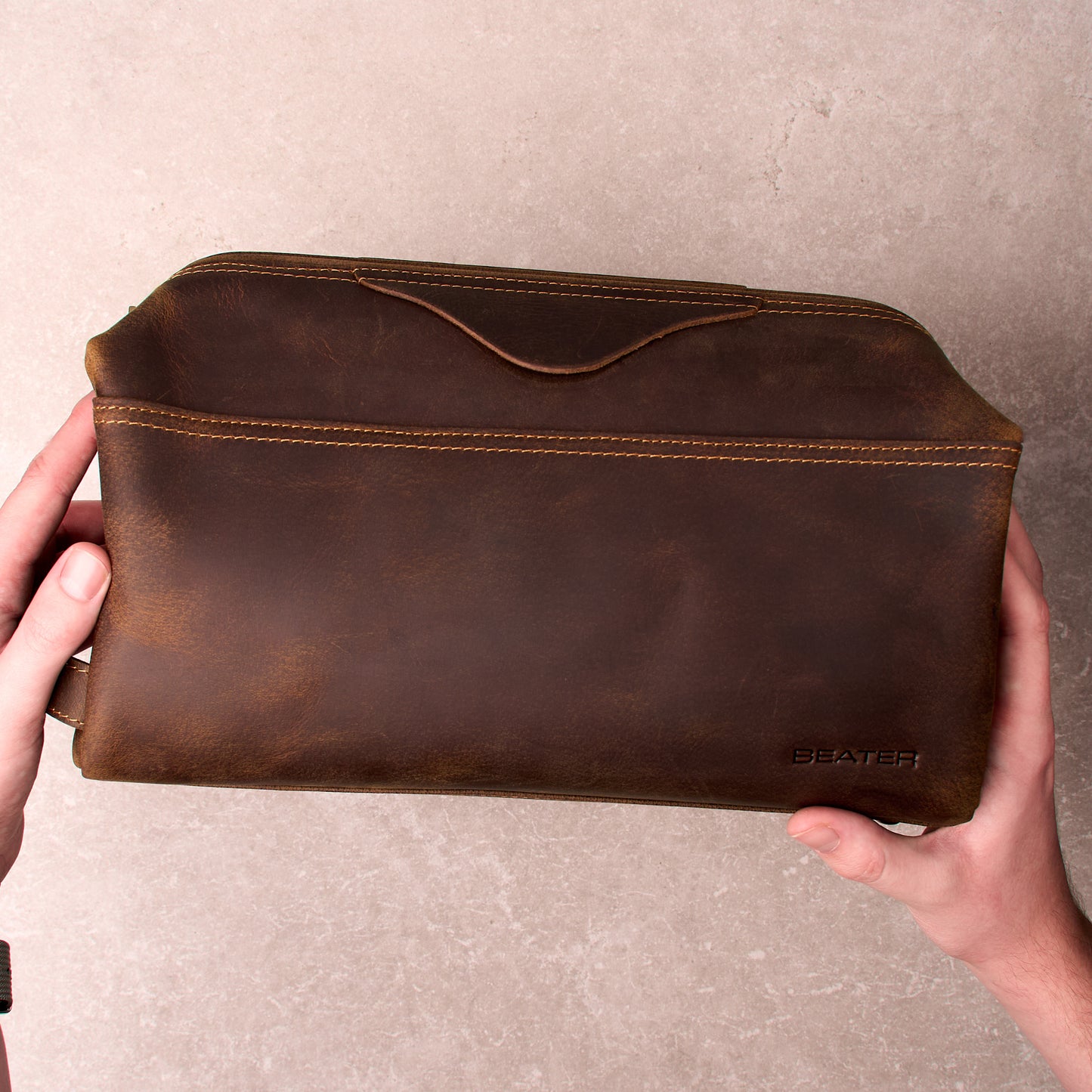 The Grafton leather toiletry bag held in two hands showing the front slip pocket and Beater logo