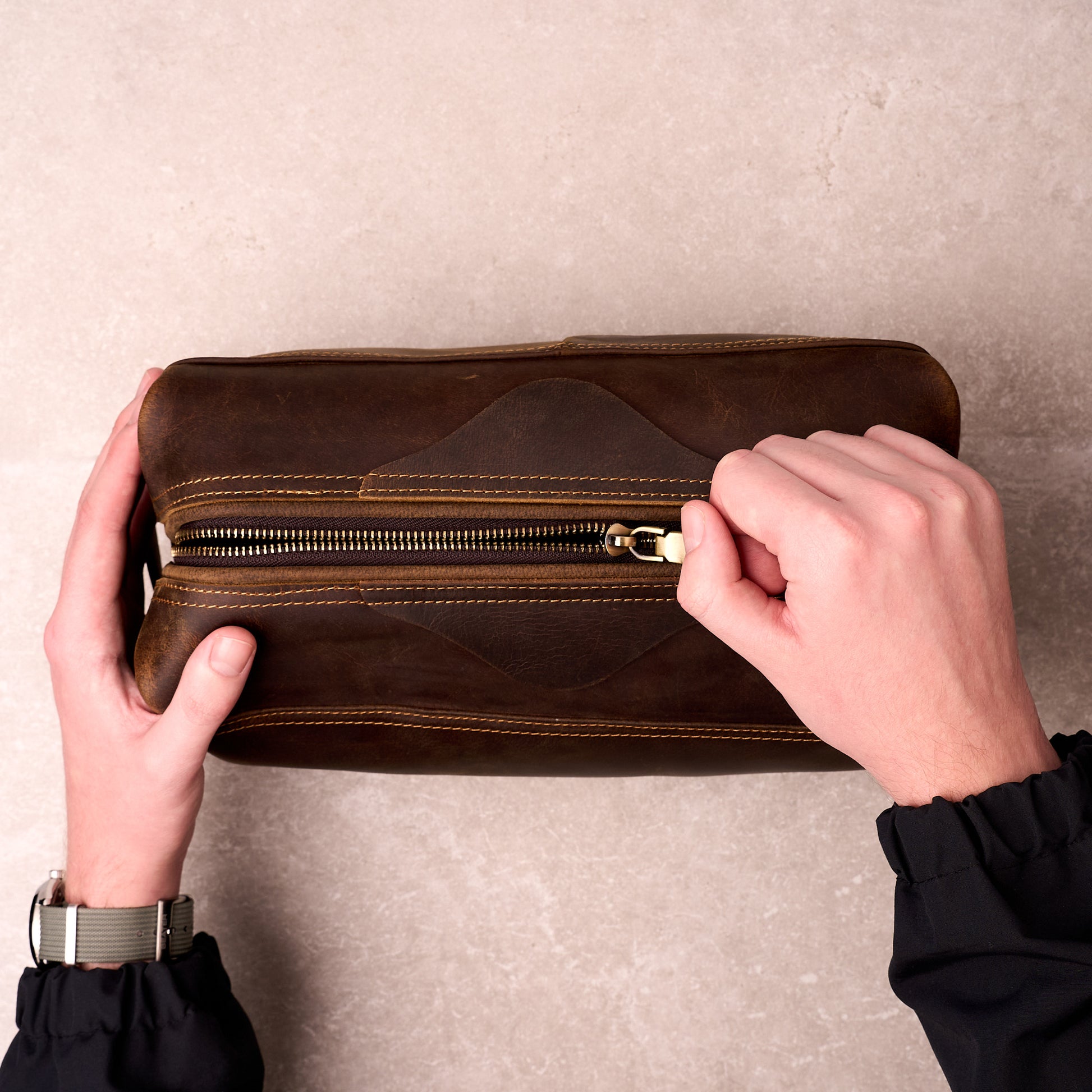 Two hands pulling open the brass zip on The Grafton leather toiletry bag